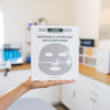 Person holding a DMK soothing and hydrating occlusive mask packaging in a blurred room setting.