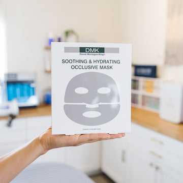 Person holding a DMK soothing and hydrating occlusive mask packaging in a blurred room setting.