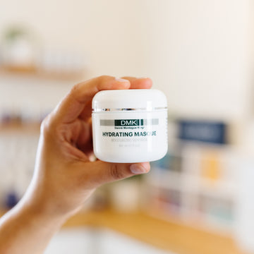Hand holding a jar of DMK Hydrating Masque in an esthetician's office.