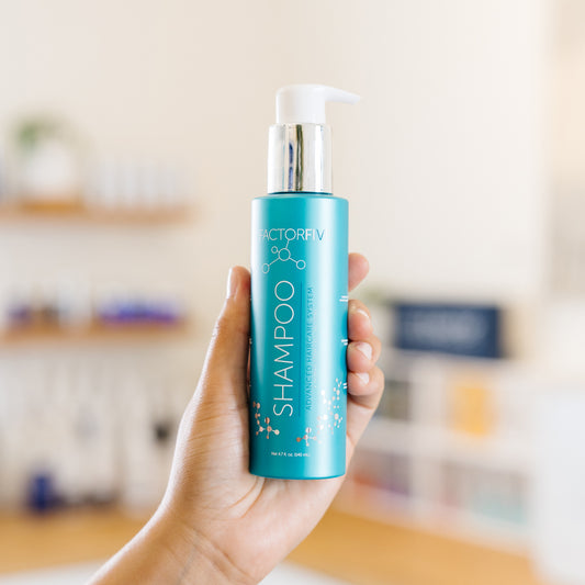 Hand holding a blue FactorFive shampoo bottle labeled 'Shampoo' in an esthetician's office.