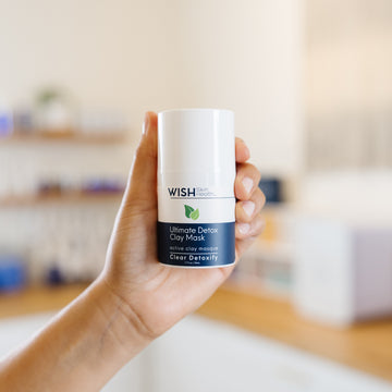 Hand holding a WISH Skin Health Ultimate Detox Clay Mask bottle in an estheticians office