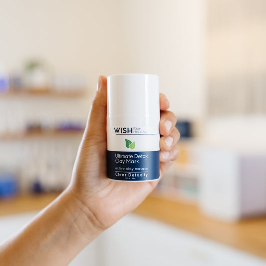 Hand holding a WISH Skin Health Ultimate Detox Clay Mask bottle in an estheticians office