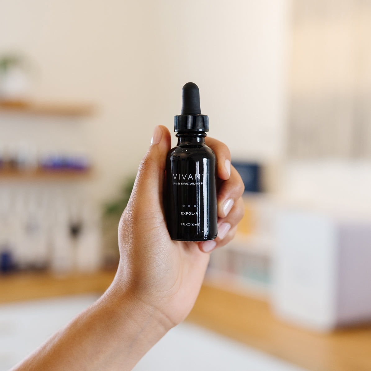 Hand holding a black bottle labeled Vivant Exfol-A with a dropper in an estheticians office
