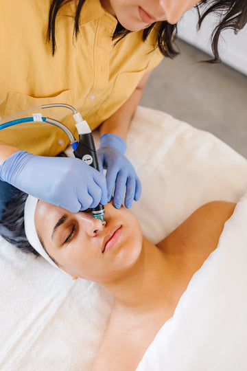 Person receiving a hydrafacial treatment with a device, wearing gloves and a headband.